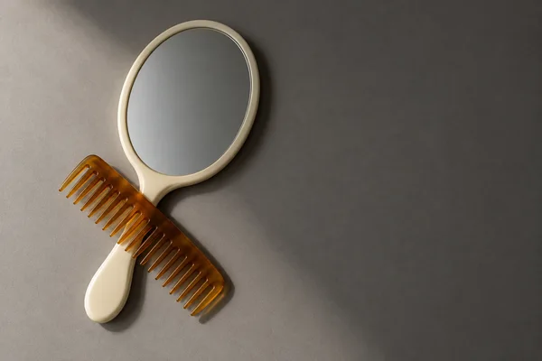 An ivory oval hand mirror and translucent amber wide-tooth comb lie on a warm-grey tabletop, with the comb crossing the mirror handle.