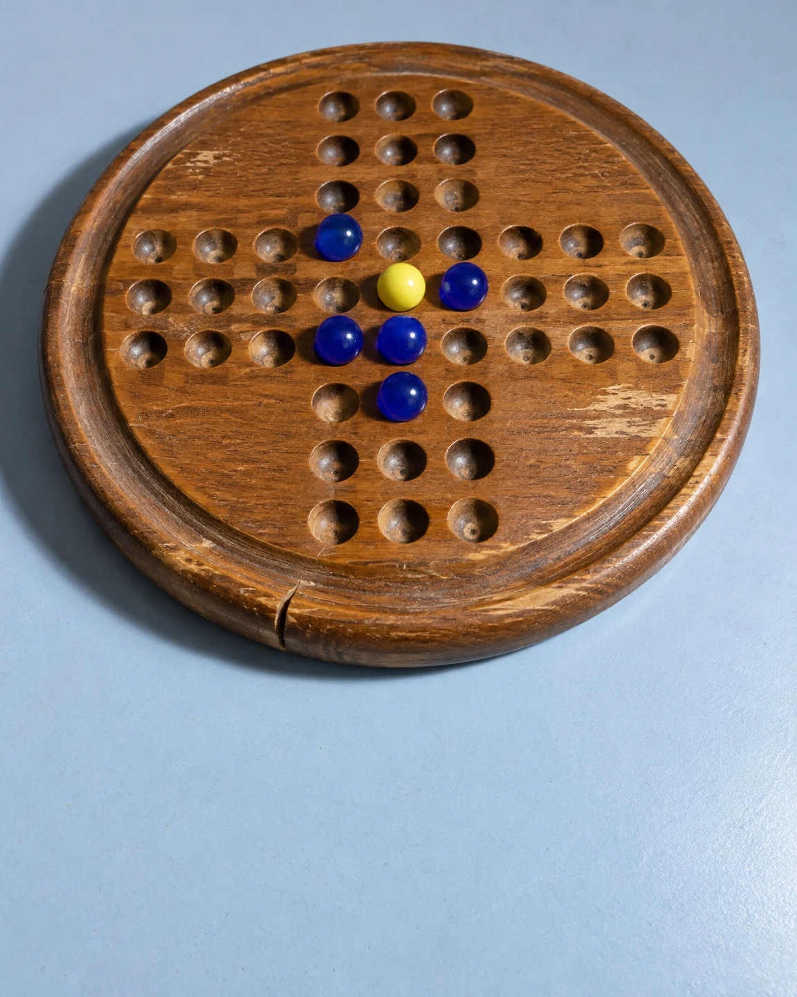 A worn circular wooden marble-solitaire board with a cross-shaped array of holes rests on a pale blue tabletop.
