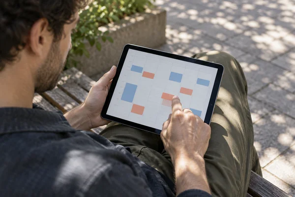 An adult viewed over the shoulder sits on a wooden courtyard bench, holding a tablet over olive trousers and touching a coral block on a pale calendar grid.