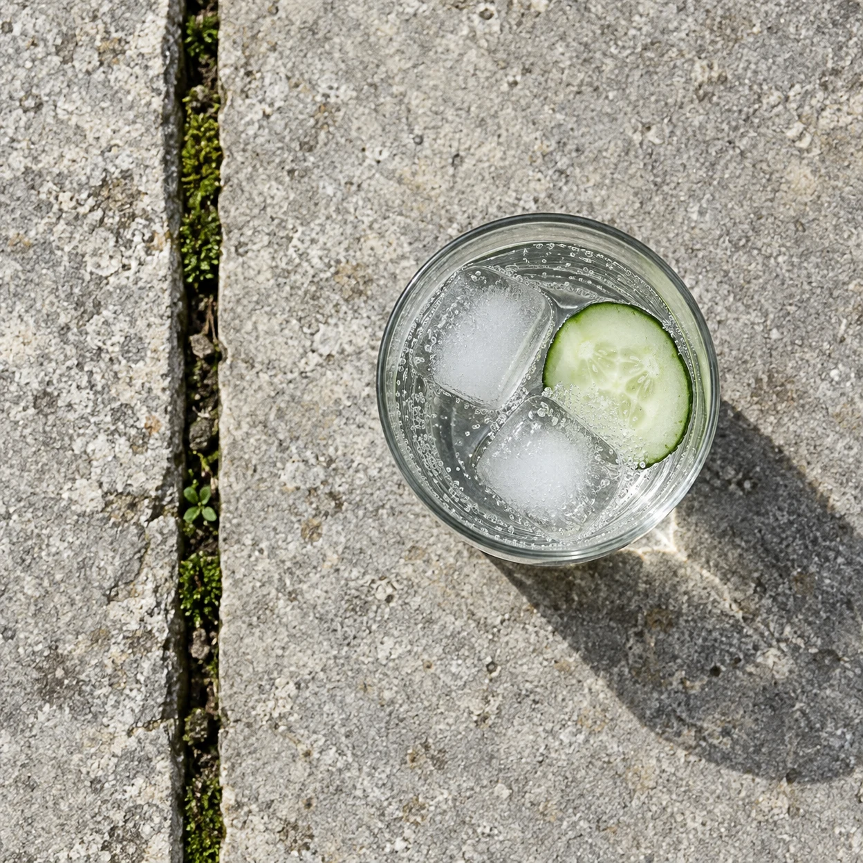 An overhead view of a clear tumbler of sparkling water with two ice cubes and a cucumber slice on a grey stone step, beside a vertical moss-filled joint.
