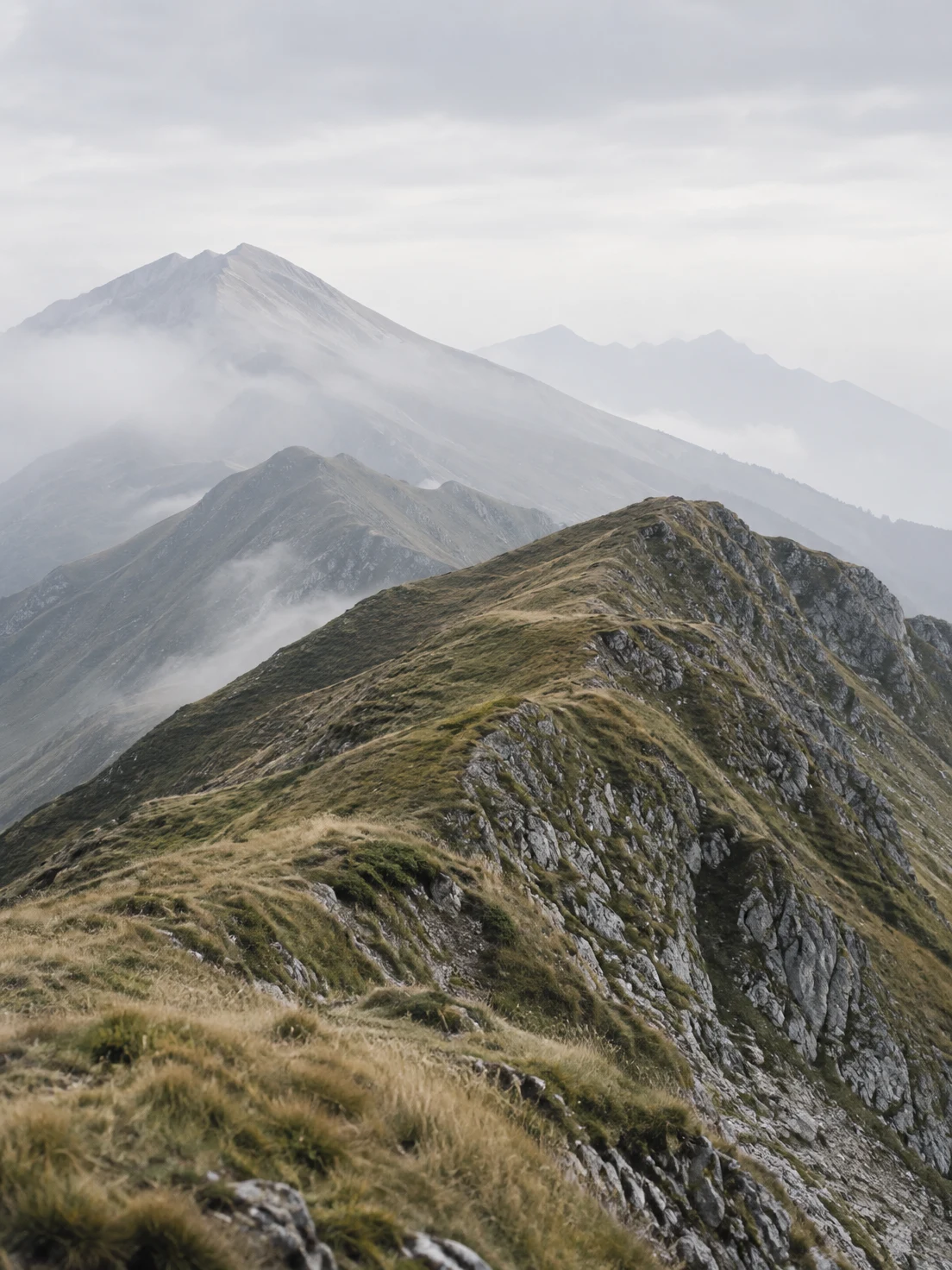 A rugged, grass-covered mountain ridge runs diagonally across the foreground, with layered grey ridges fading into mist beneath a pale overcast sky.