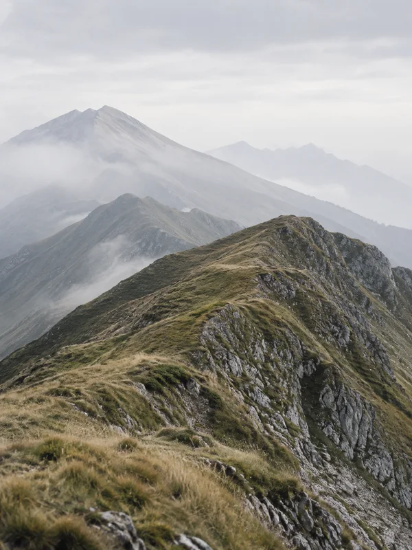 A rugged, grass-covered mountain ridge runs diagonally across the foreground, with layered grey ridges fading into mist beneath a pale overcast sky.