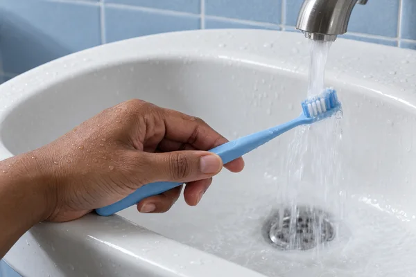 A medium-brown hand holds a powder-blue toothbrush beneath a brushed-steel faucet over a white porcelain sink, with pale-blue tiles behind it and water running…