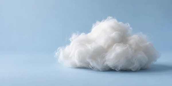 A fluffy white wool cloud sculpture rests on a pale-blue surface against a seamless blue backdrop, with open space to the left.