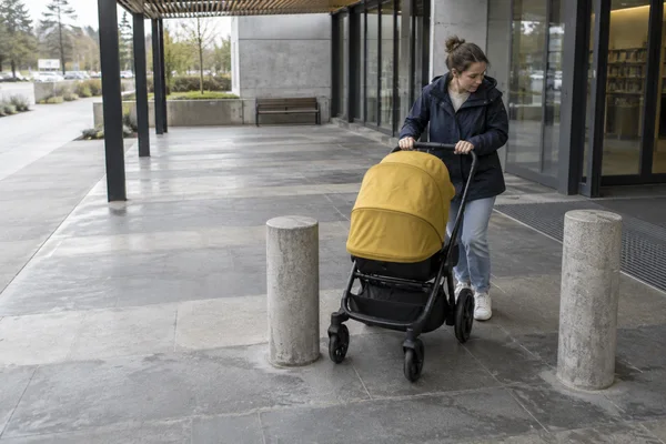 A person in a navy rain jacket and faded jeans steers a mustard-yellow stroller between two concrete bollards outside a glass-fronted public building, with…