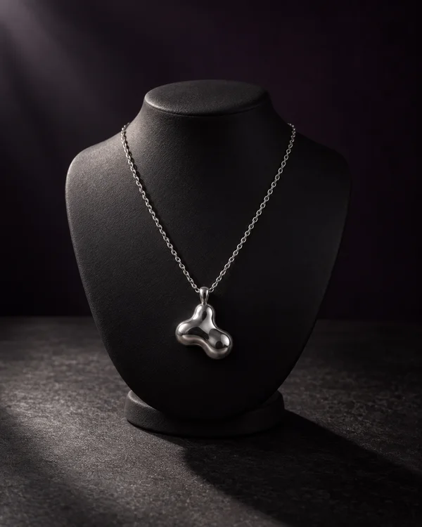 A polished silver necklace with a rounded three-lobed pendant hangs from a textured charcoal-black display bust on a dark tabletop, lit from the upper left…