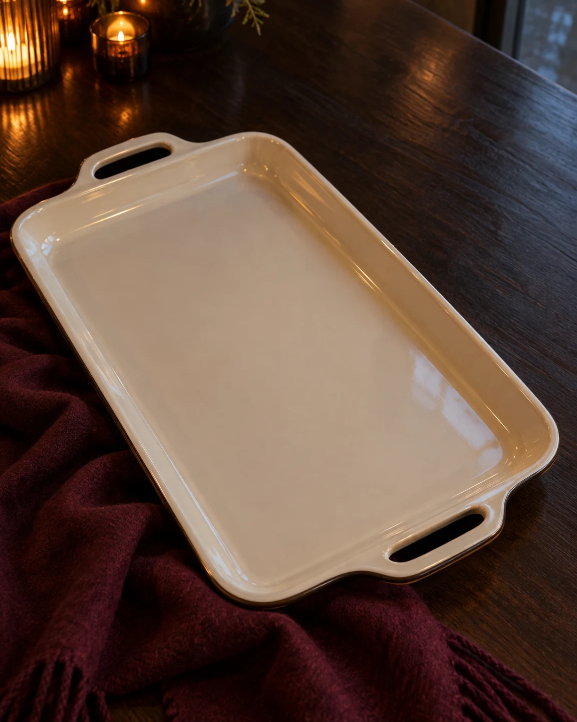 An empty cream enamel serving tray with integrated handles lies diagonally across a dark wooden table, overlapping a burgundy wool scarf.