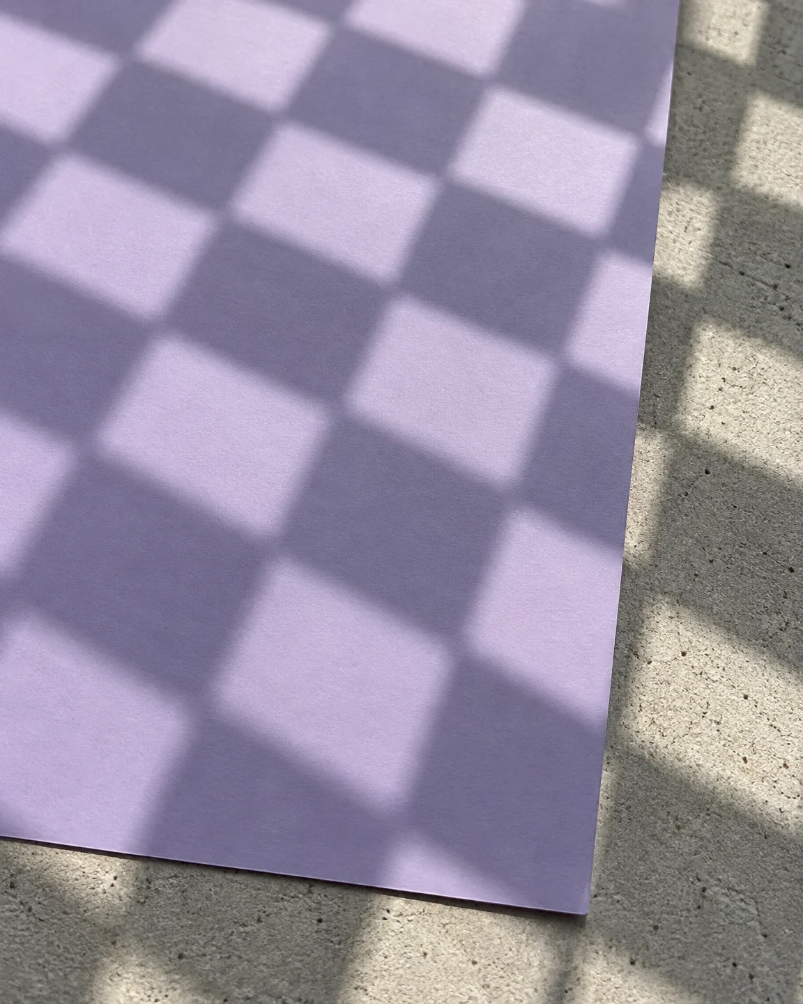 A large lilac paper sheet lies on pale, speckled concrete, with a grid of alternating light and shaded squares crossing both surfaces.