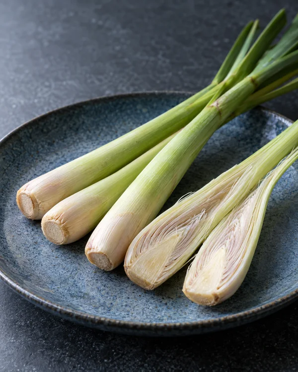 Four pale-green lemongrass stalks lie on a shallow blue glazed plate over a dark charcoal worktop; one is split lengthwise at the base to show its ivory…