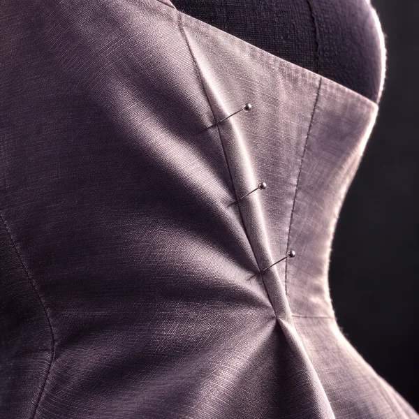 A close-up of rose-violet fabric stretched over a curved dark dress form, with a raised diagonal fold held by three silver pins.