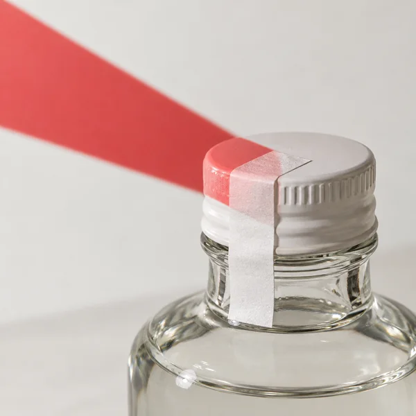 Close-up of a clear glass bottle with a white ribbed screw cap and a white paper seal running over the cap and down the neck; a red diagonal light projection…