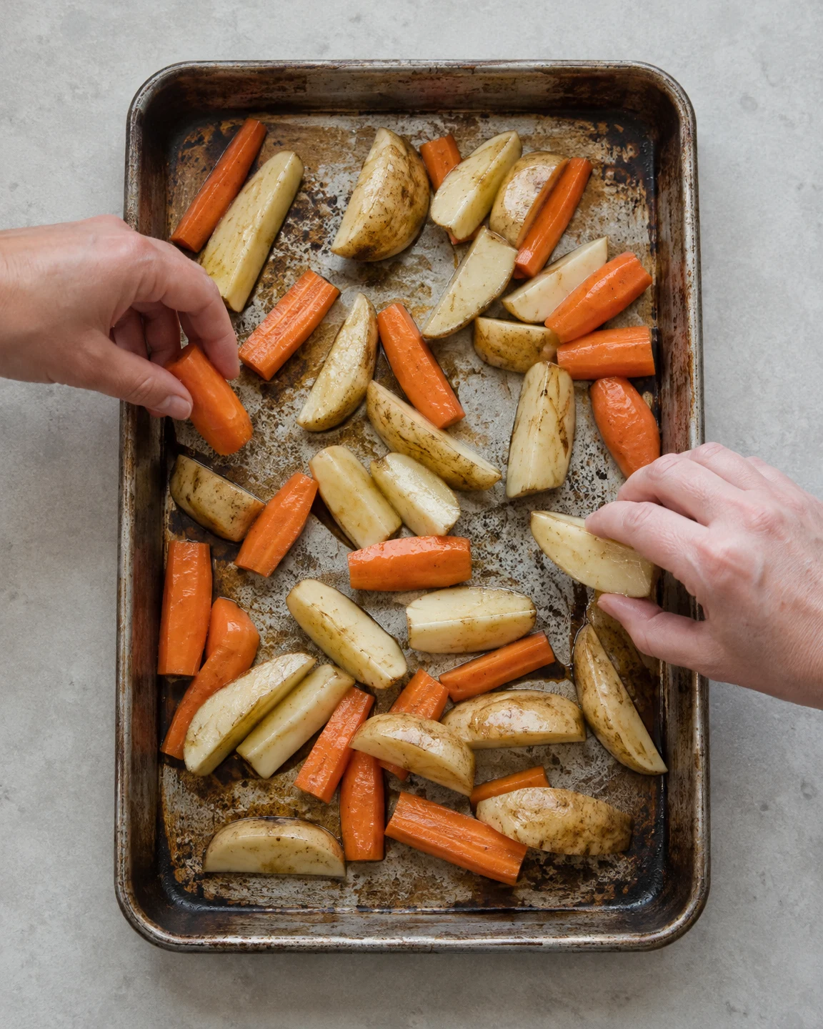Overhead view of a heavily worn rectangular roasting tin filled with potato wedges and carrot pieces on a pale-grey counter, with a hand entering from each…