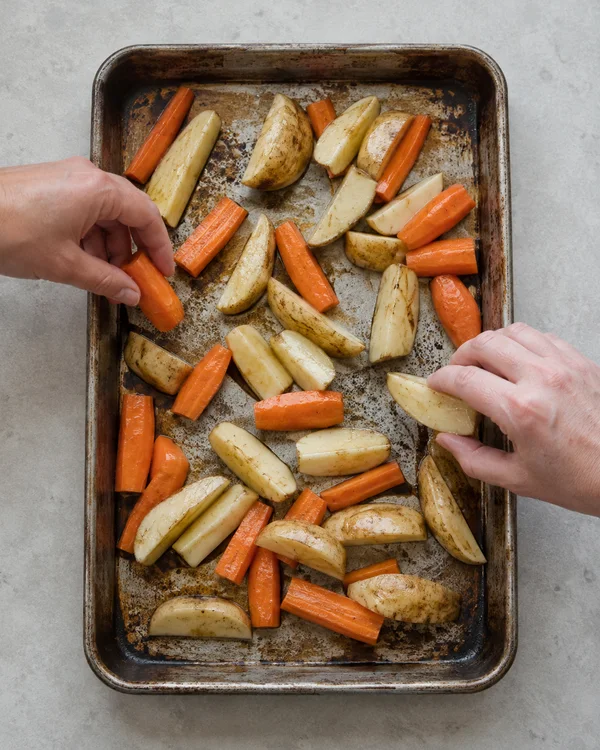 Overhead view of a heavily worn rectangular roasting tin filled with potato wedges and carrot pieces on a pale-grey counter, with a hand entering from each…