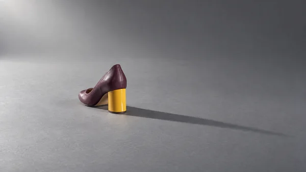 A plum leather pump with a glossy yellow block heel stands on a gray studio floor, casting a long shadow to the right.