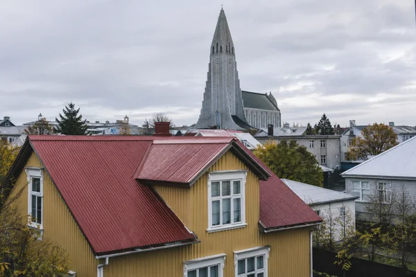 An ochre house with white-trimmed windows and a red corrugated roof fills the foreground, with Hallgrímskirkja rising above Reykjavík rooftops beneath a cloudy…