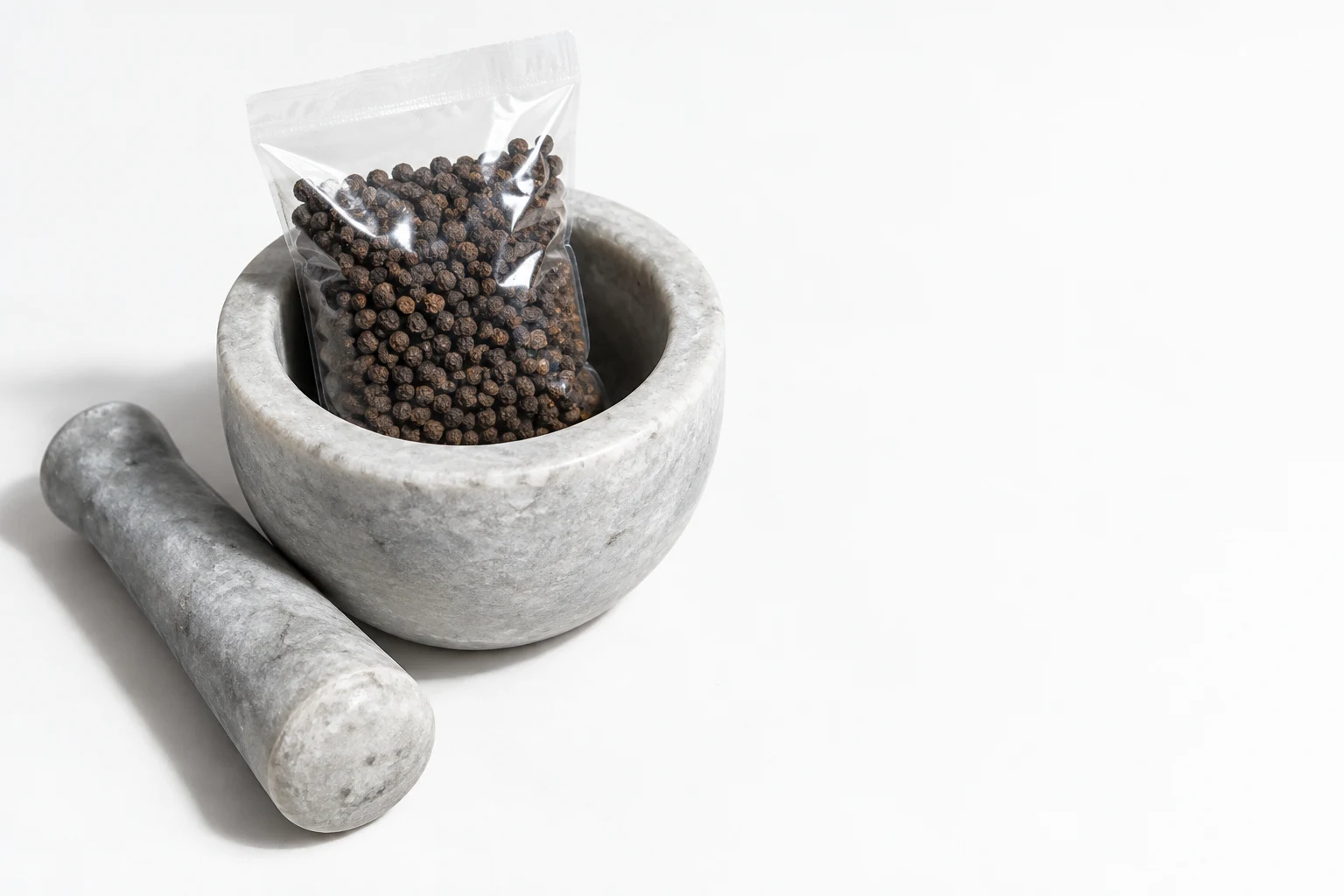 A clear sealed plastic pouch filled with black peppercorns stands inside a pale grey stone mortar, with a matching stone pestle lying diagonally beside it on a…