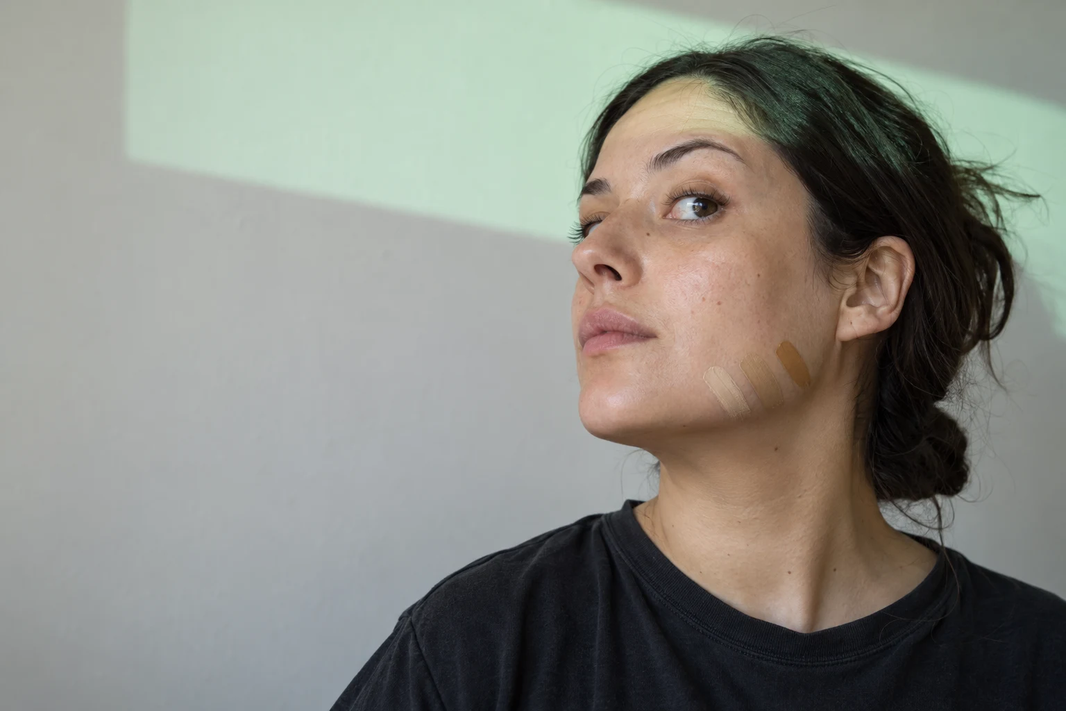 A woman in a dark T-shirt turns her chin upward and looks off-frame, with three foundation swatches along her jaw and a mint-green rectangular band of light…