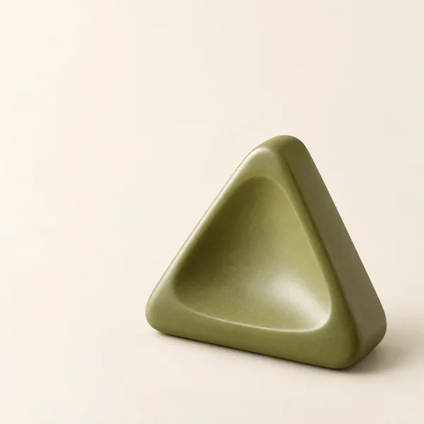 A thick olive-green triangular ceramic form with rounded corners and a scooped front face rests against a seamless warm-cream background, with ample open space…