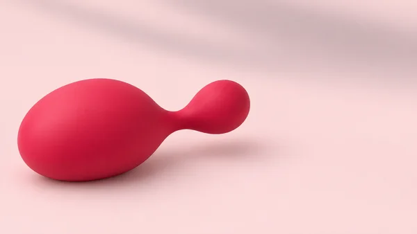 A smooth raspberry-red, two-lobed sculptural form rests on a pale pink seamless surface, with a soft shadow beneath it and open space to the right.