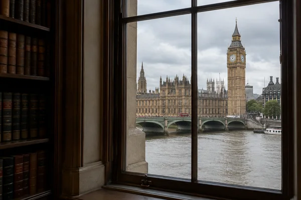 A view across the Thames to Elizabeth Tower and the Palace of Westminster, framed by a tall window inside a room with dark wooden bookshelves.