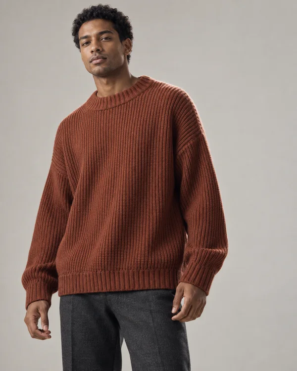 A male model with short curls wears an oversized rust-brown ribbed sweater and charcoal trousers against a pale warm-grey studio background, with open space to…