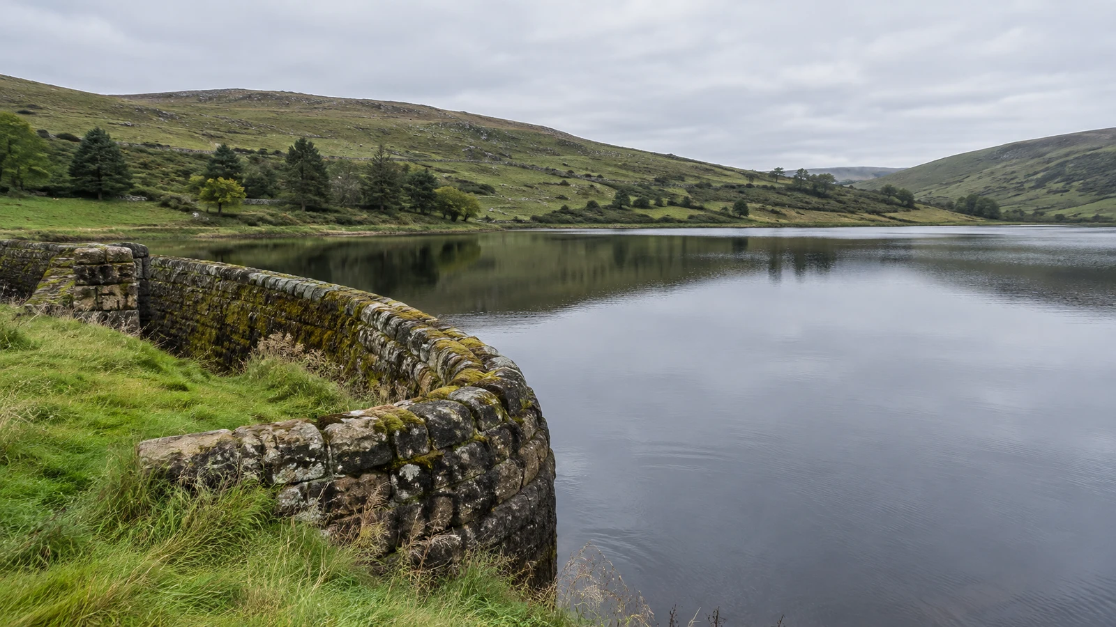 A moss-covered curved stone wall borders a calm upland reservoir, with green hills and scattered trees reflected in the water beneath an overcast sky.