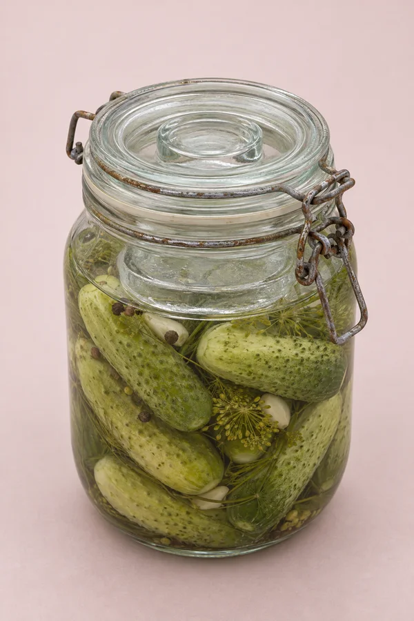 A large vintage glass jar of pickled cucumbers stands against a pale blush seamless background.