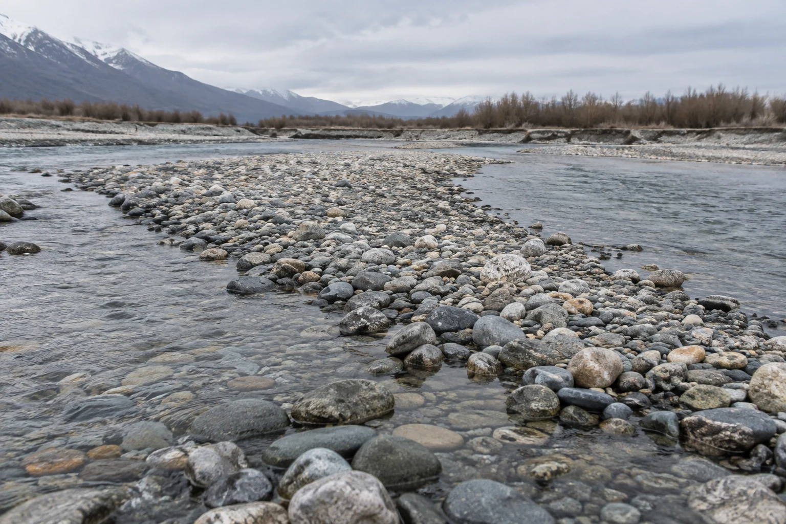 A shallow braided river flows around a diagonal bar of rounded grey, brown and ochre stones, with scrub along the far bank and snow-covered mountains beneath…