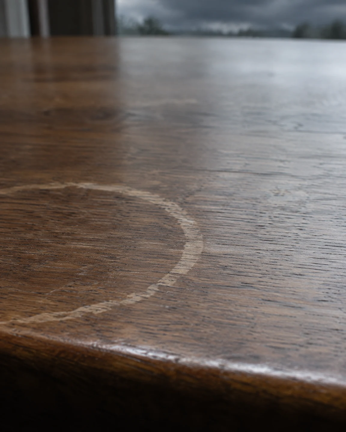 A large pale cup ring crosses the lower-left area of a brown wooden tabletop, with the rounded front edge at the bottom and a softly blurred cool-toned…
