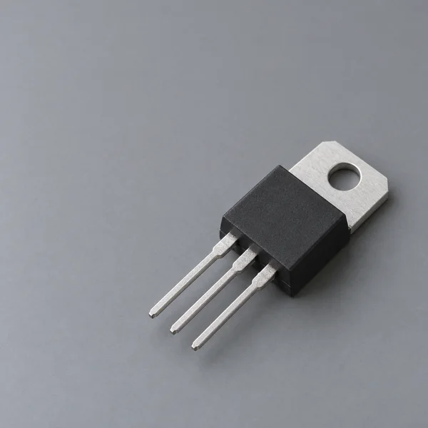 A black three-lead power transistor lies diagonally on a cool gray surface, with its silver mounting tab and circular hole visible behind the body.