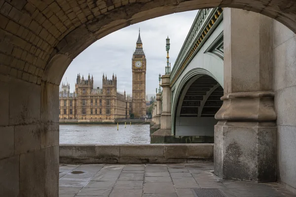 A stone-vaulted opening frames Elizabeth Tower and the Palace of Westminster across the grey Thames, with Westminster Bridge filling the right side and…