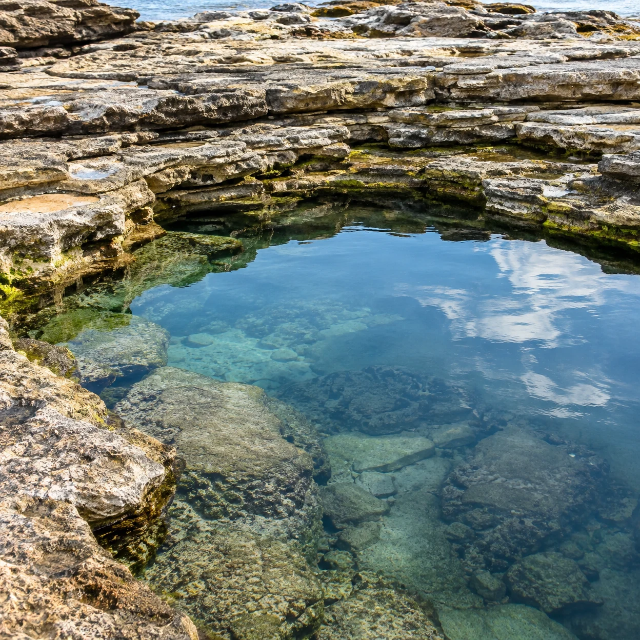 A clear blue coastal rock pool sits among layered, pale limestone ledges, with submerged stones visible beneath sky reflections and open water beyond the shelf.