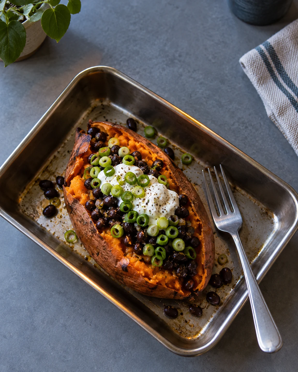 A split roasted sweet potato topped with black beans, white yogurt and sliced scallions sits in a diagonal stainless-steel roasting tin, with a fork beside it.