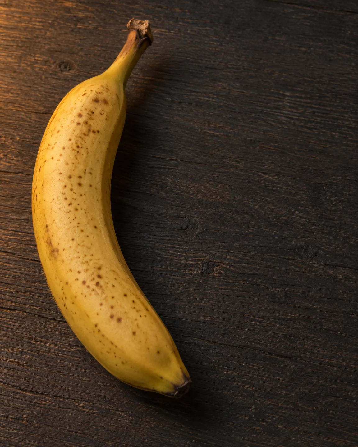 A freckled yellow banana lies vertically along the left side of a dark wooden tabletop, with warm light falling across the peel and open space to the right.