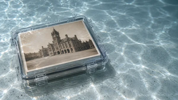 A clear rectangular hard case rests on a pale blue-grey floor, with a sepia photograph of a stone town hall visible inside; rippled light and small bubbles…