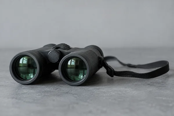 A pair of black binoculars rests on a cool-grey tabletop, with green-tinted objective lenses reflecting a window and a woven strap trailing to the right.