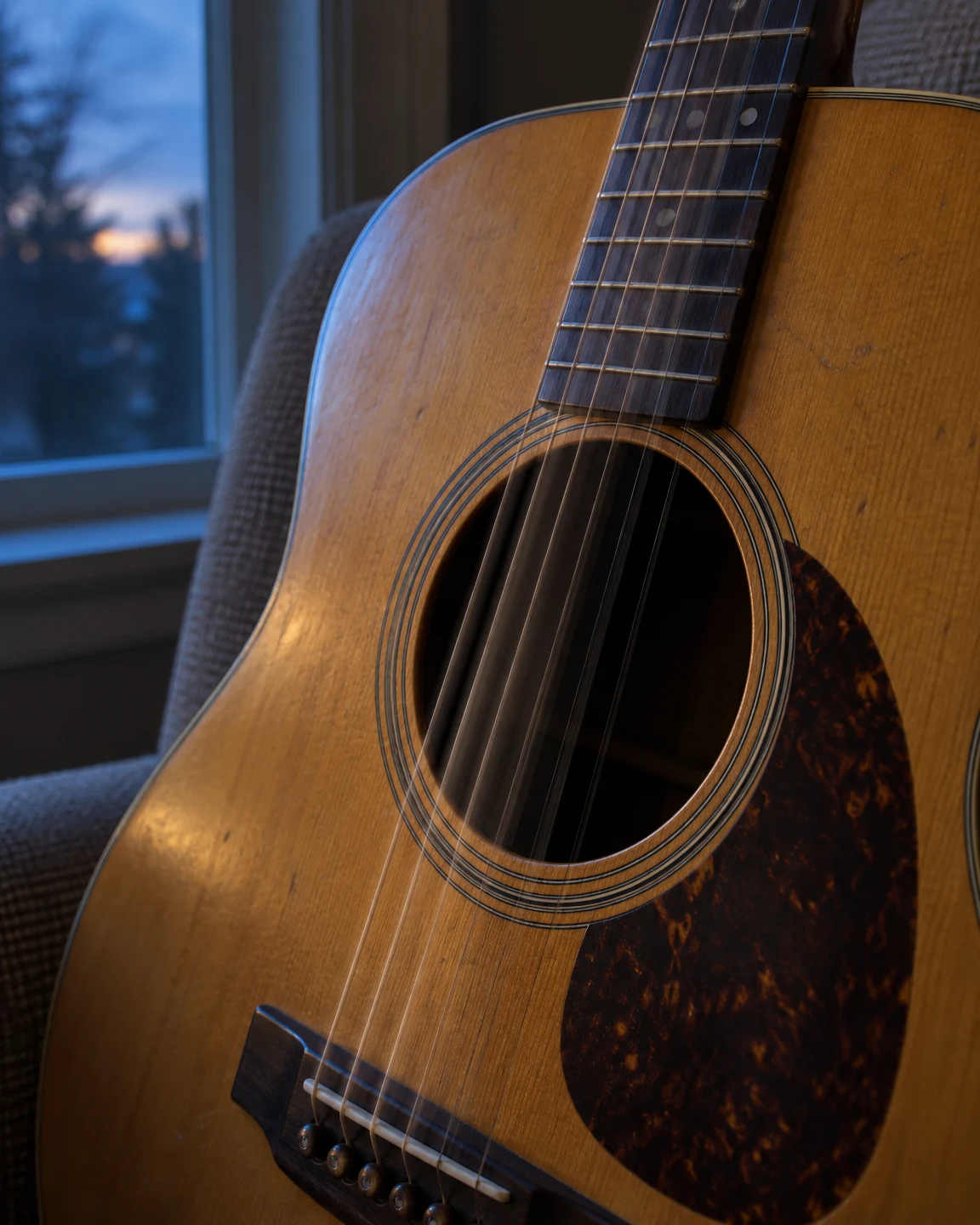 Close oblique view of a honey-coloured acoustic guitar leaning against a muted upholstered chair beside a blue-hour window, with the dark sound hole and…