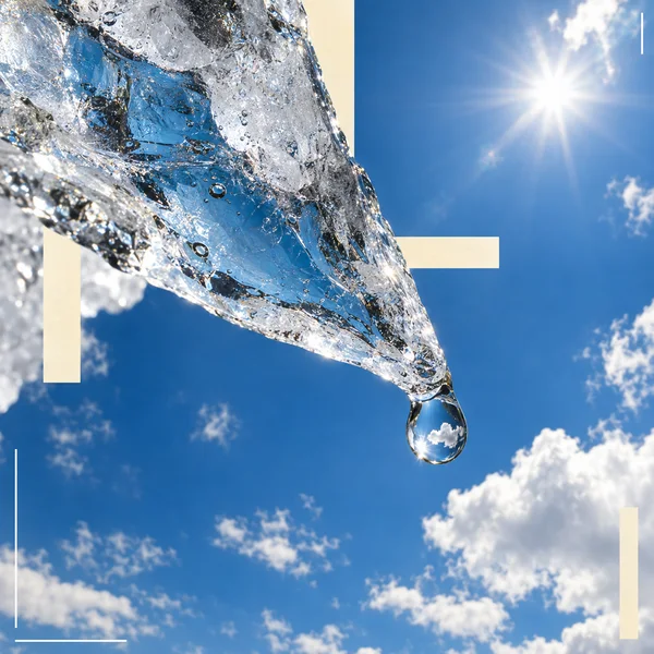 A large transparent, faceted ice shard crosses diagonally from the upper left toward a hanging droplet against a blue sky with scattered clouds and a bright…