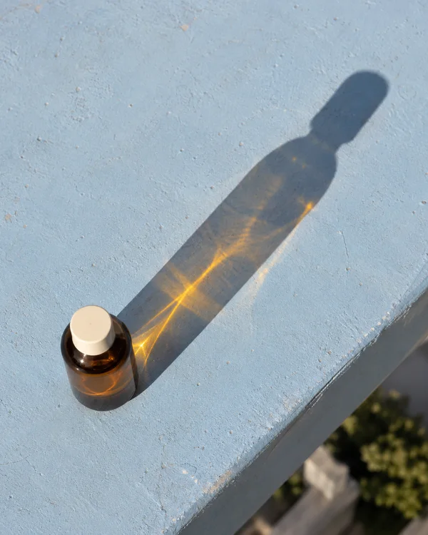 A squat amber glass bottle with a plain cream cap stands on a pale-blue painted concrete ledge, casting a long shadow toward the upper right with a warm golden…