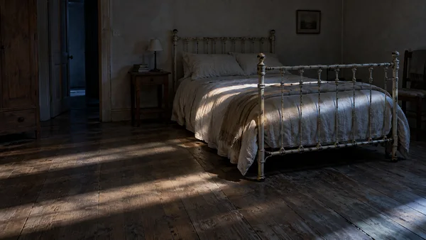 A dim, weathered bedroom with a chipped cream iron bed and rumpled pale linen on the right, scuffed timber floor across the foreground, and an open doorway to…