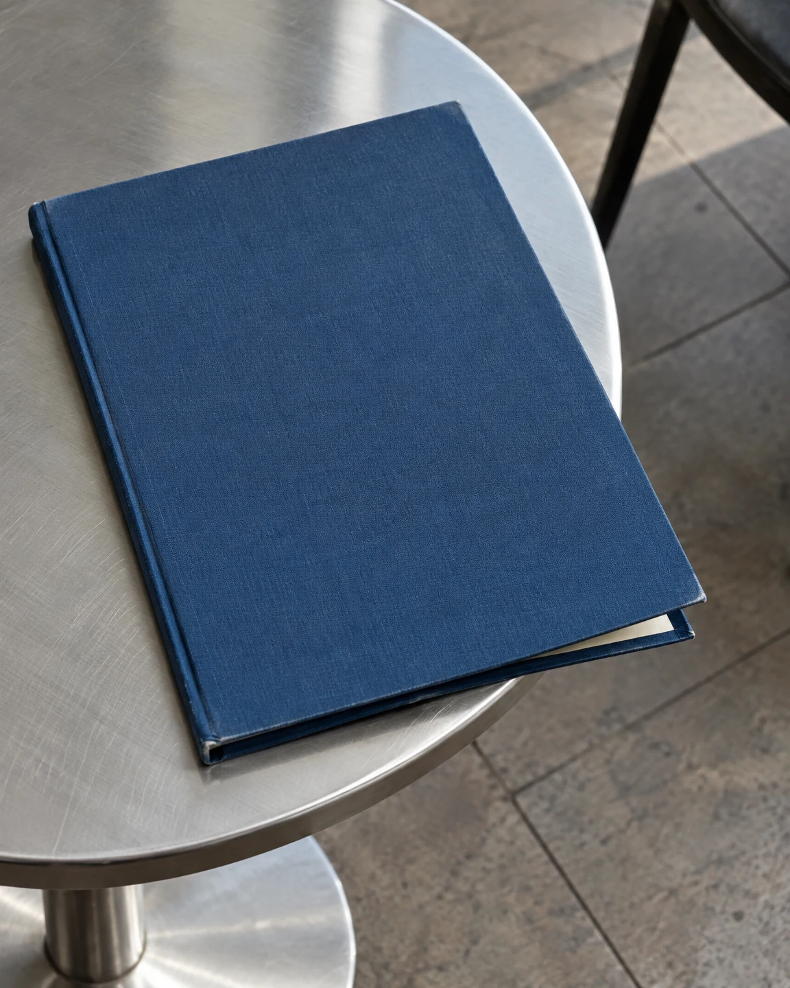 A plain dark-blue cloth menu binder lies diagonally on a brushed-aluminium café table, with a thin cream strip visible beneath its slightly lifted lower corner.
