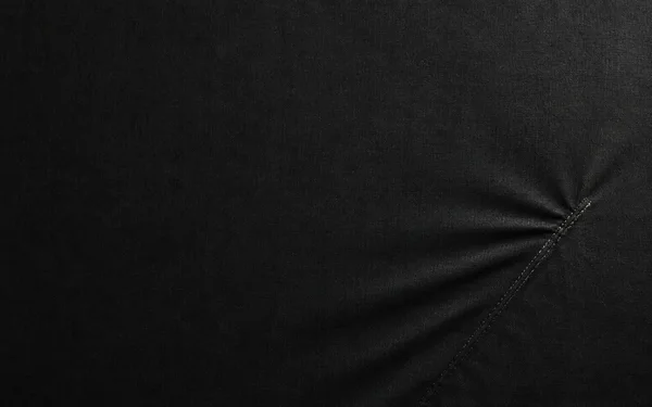 A close, straight-on view of dark woven fabric fills the frame, with a stitched diagonal seam in the lower-right area and shallow creases radiating across the…