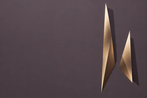 Two unequal, sharply tapered champagne-bronze planes with dark offset shadow shapes sit on the right side of a broad plum-grey field, leaving extensive empty…