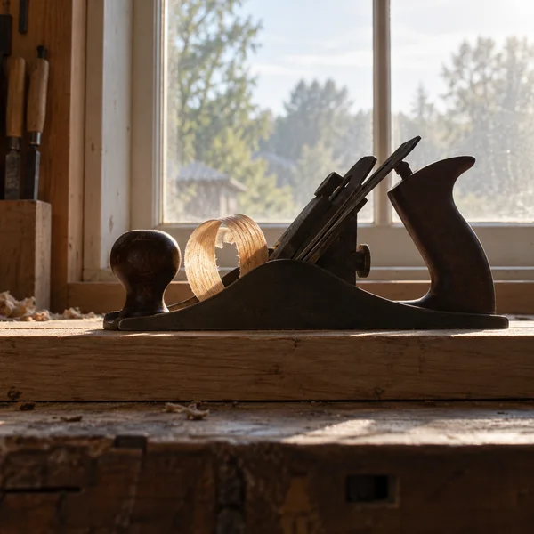 A dark metal hand plane rests on a timber workbench in front of a bright workshop window, with a honey-gold wood shaving curled beside it and other tools…
