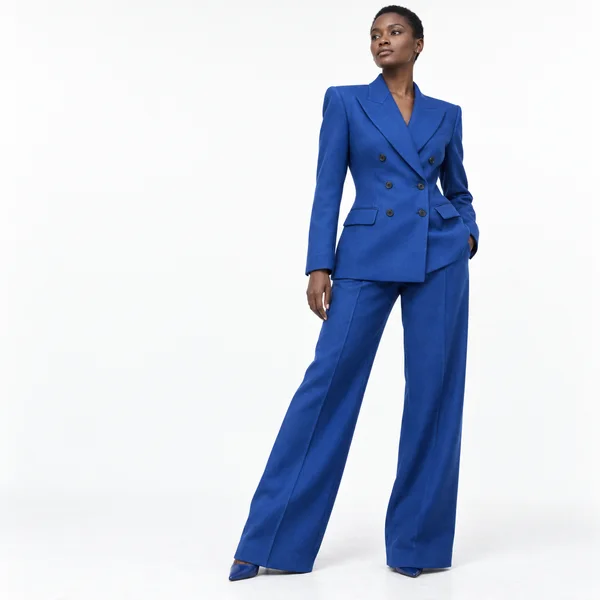 A woman with closely cropped hair stands right of center against a bright white studio backdrop, wearing a cobalt-blue double-breasted suit with wide-leg…