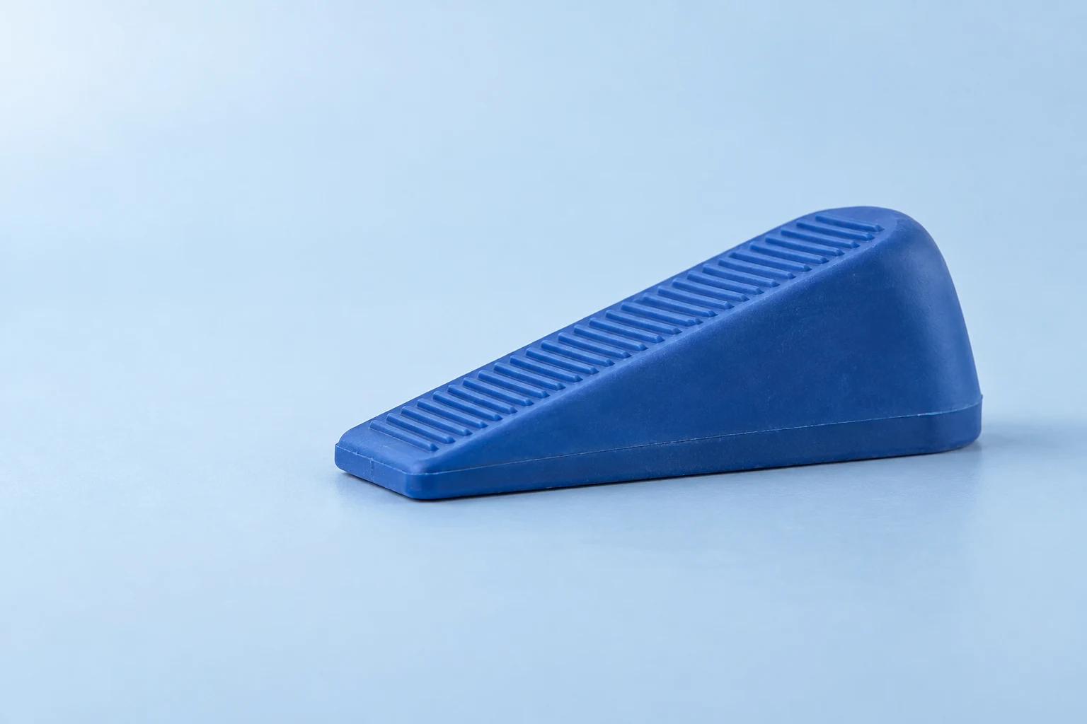 A cobalt-blue rubber doorstop rests on a pale-blue tabletop, shown from a low three-quarter angle with its ribbed top and broad triangular side visible.