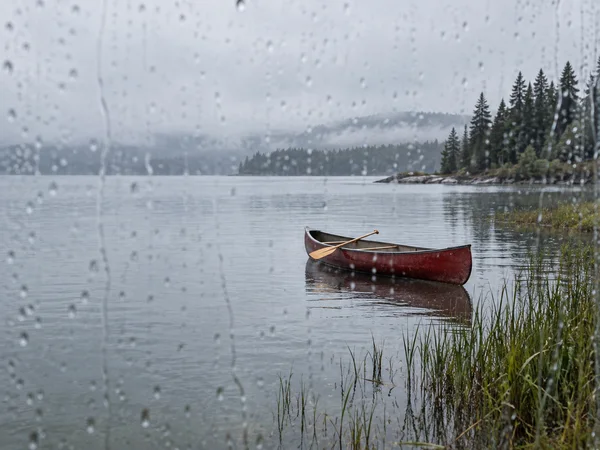 An empty red canoe with a paddle resting across it floats on a grey-green lake, seen through a rain-speckled window.