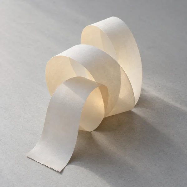A long, blank ivory paper strip curls into several overlapping upright loops on a pale-gray textured surface, with a finely serrated end in the foreground and…