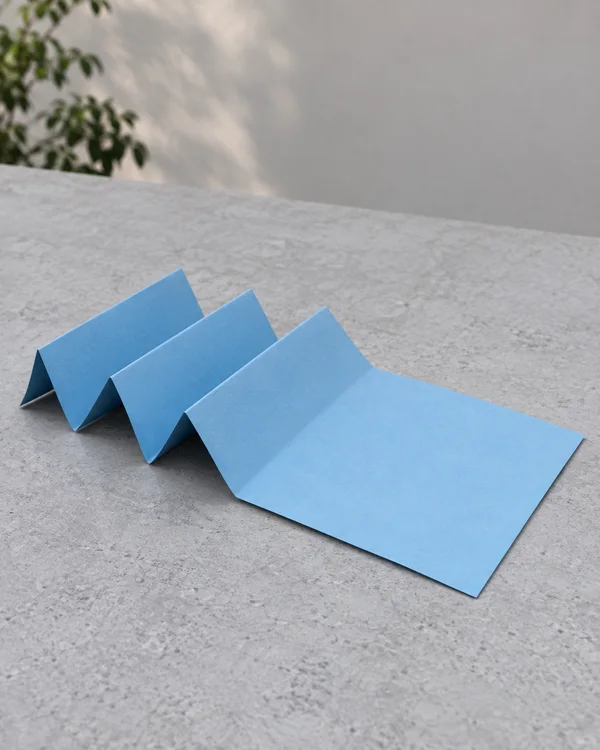A sky-blue sheet of paper folded into three raised accordion pleats and a broad flat panel rests on a textured pale-grey tabletop.