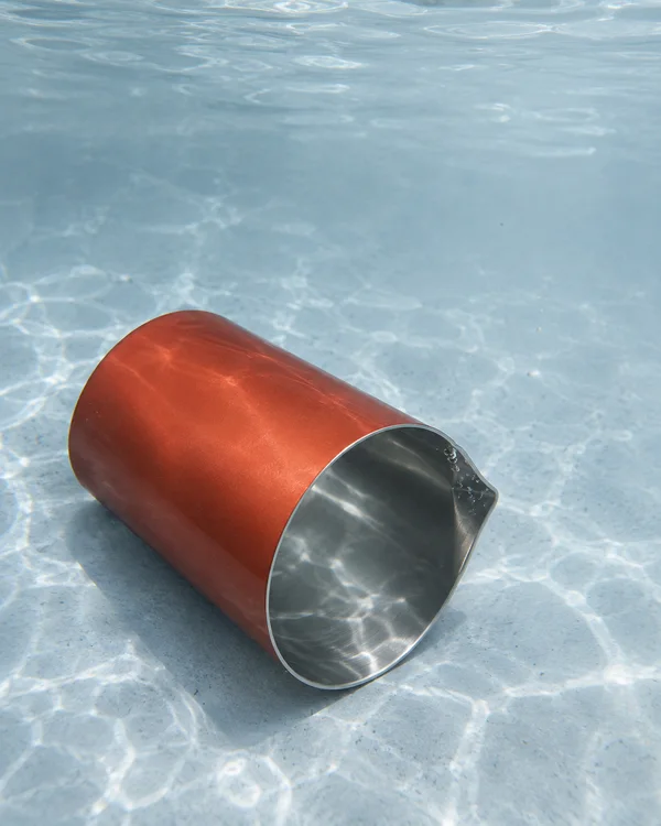 A vermilion, handle-free metal cocktail beaker lies diagonally on the pale blue-grey floor of a water tank, its silver interior and pinched pouring lip visible…